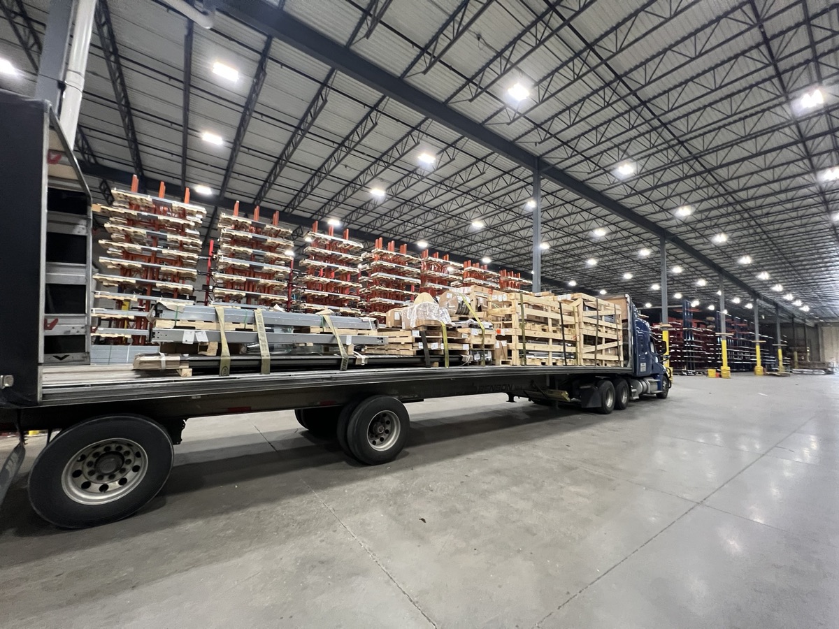 A flatbed trailer loaded indoors with steel sections and crated freight, parked inside a warehouse.